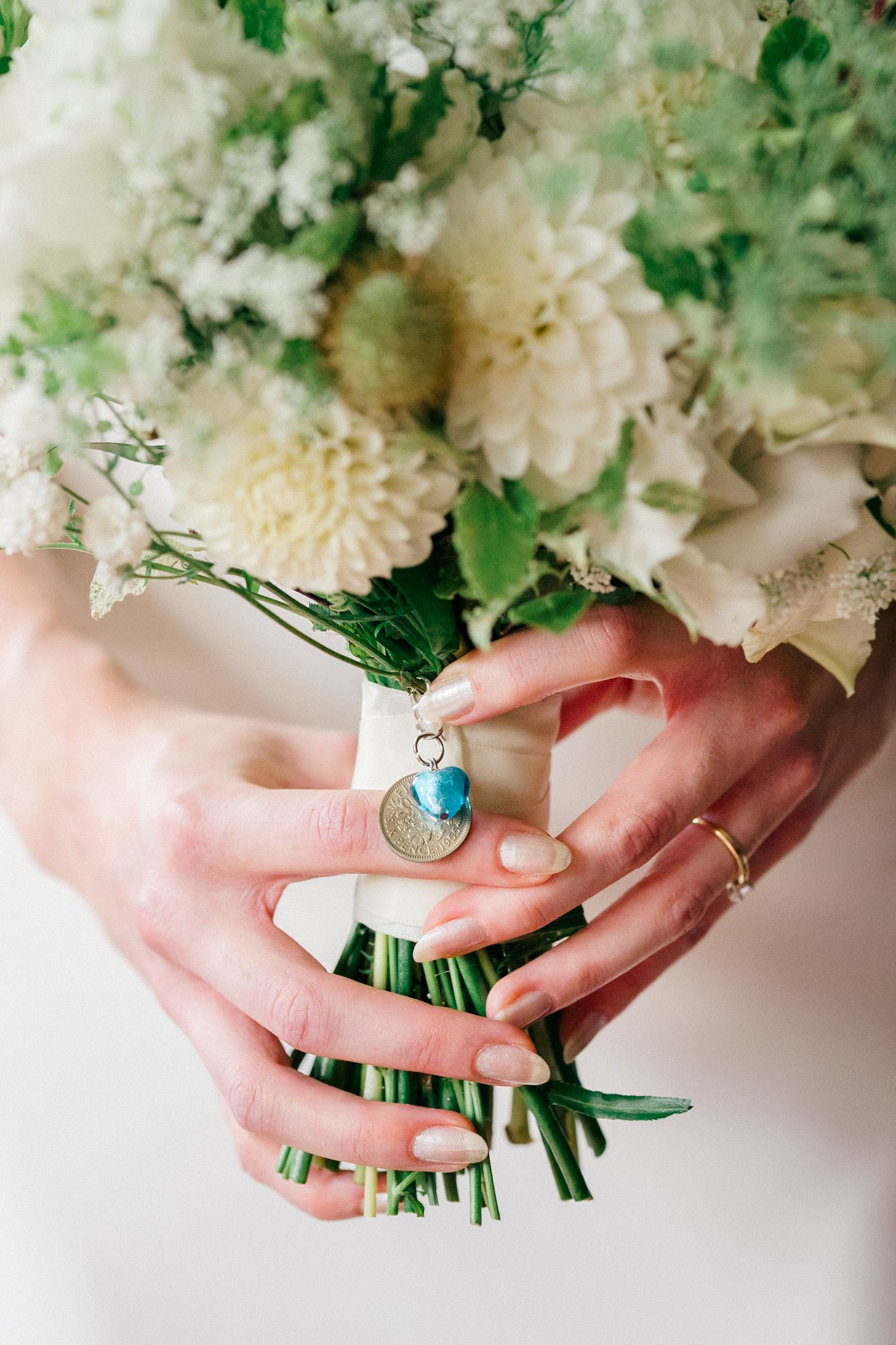 Bride holding white floral wedding bouquet with blue charm pendant and gold coin, wearing delicate rings.
