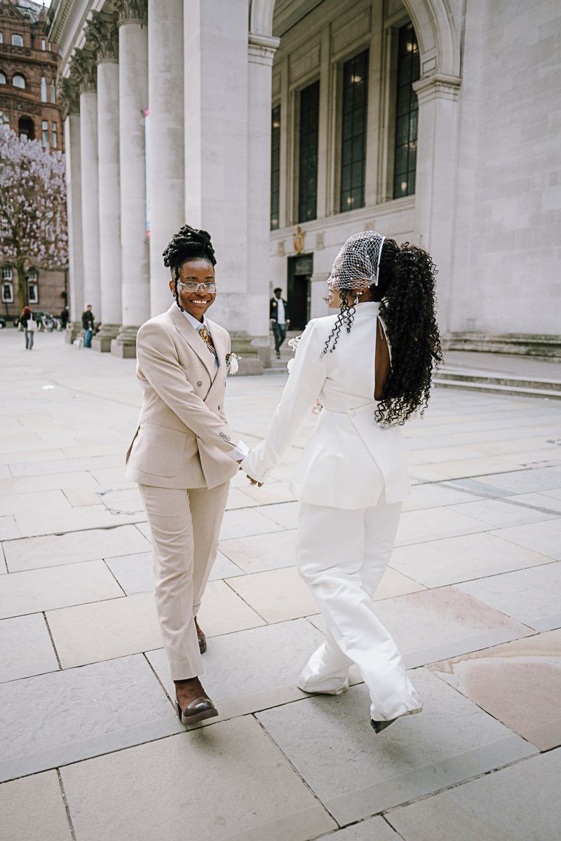 Two Black women in elegant white wedding attire hold hands and smile outside a grand building.