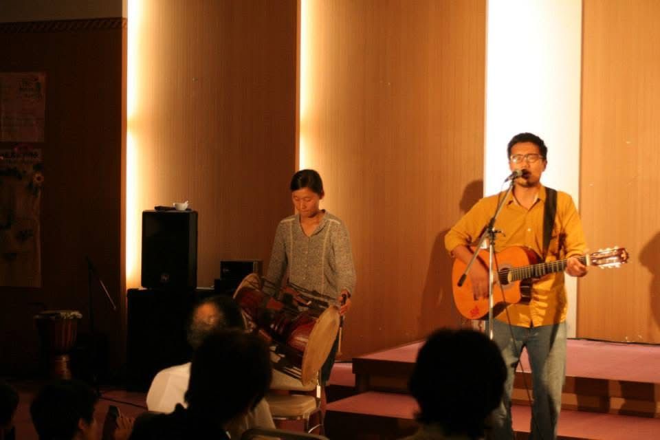 Two musicians performing live, one playing acoustic guitar and singing, with an audience watching in a warmly lit venue.