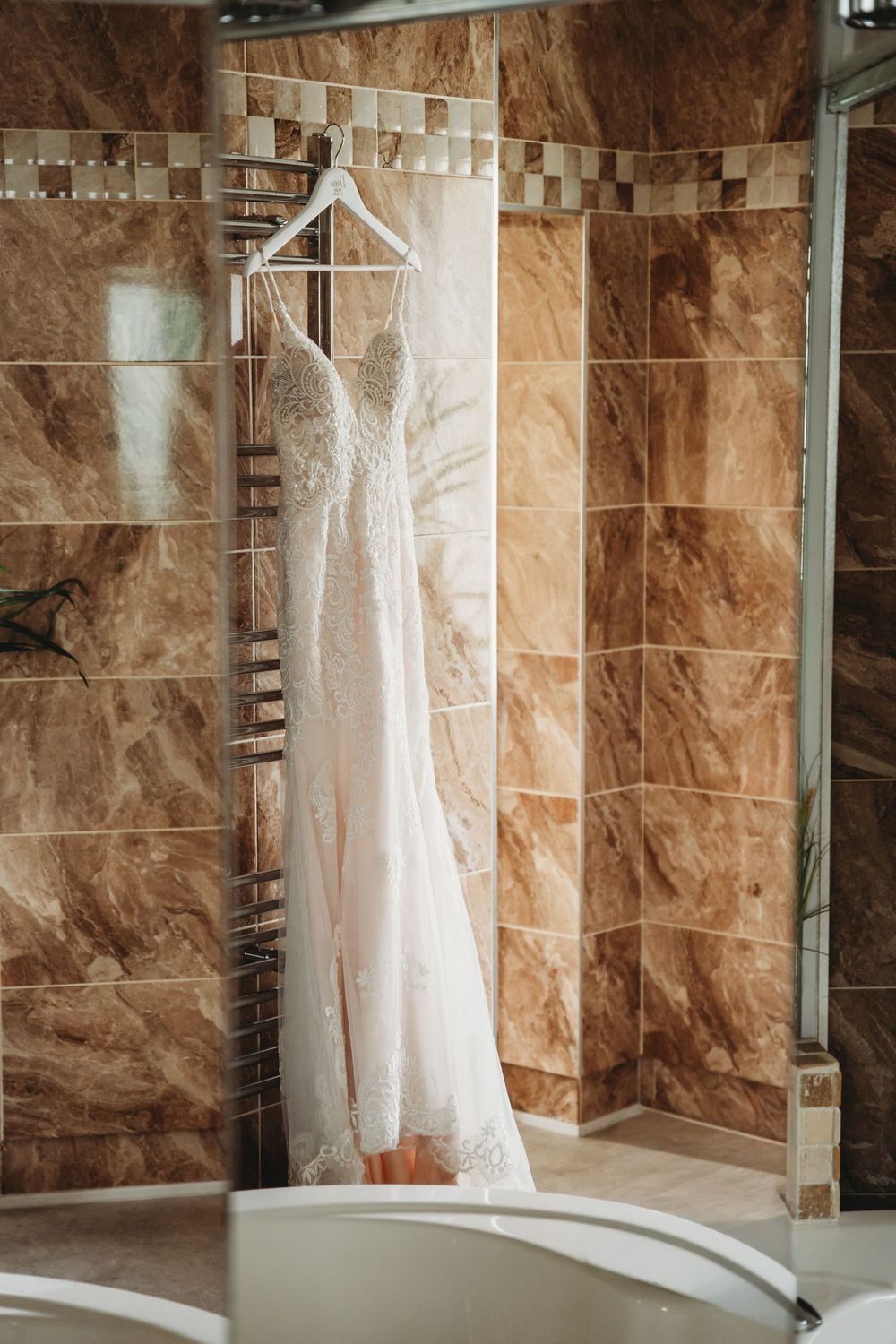 A lace wedding dress hanging on a white hanger against warm brown marble bathroom tiles.