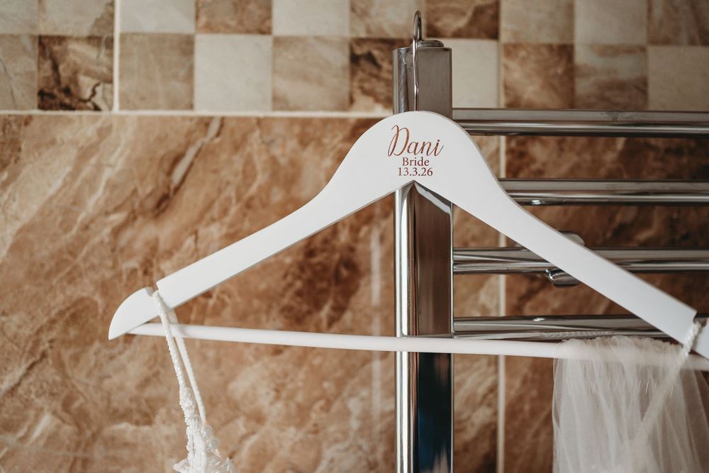 Personalized white wooden wedding hanger with bride's name displayed on a chrome towel rail in a tiled bathroom.
