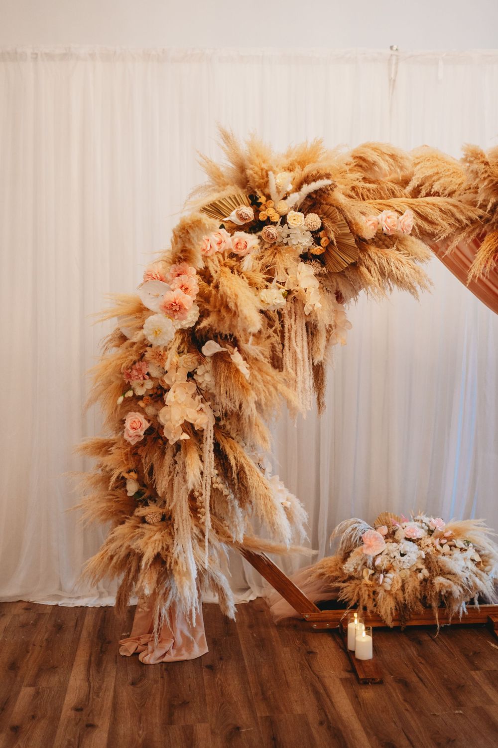 Boho wedding arch adorned with pampas grass, dried flowers, and blush roses on wooden floor with white drape backdrop.