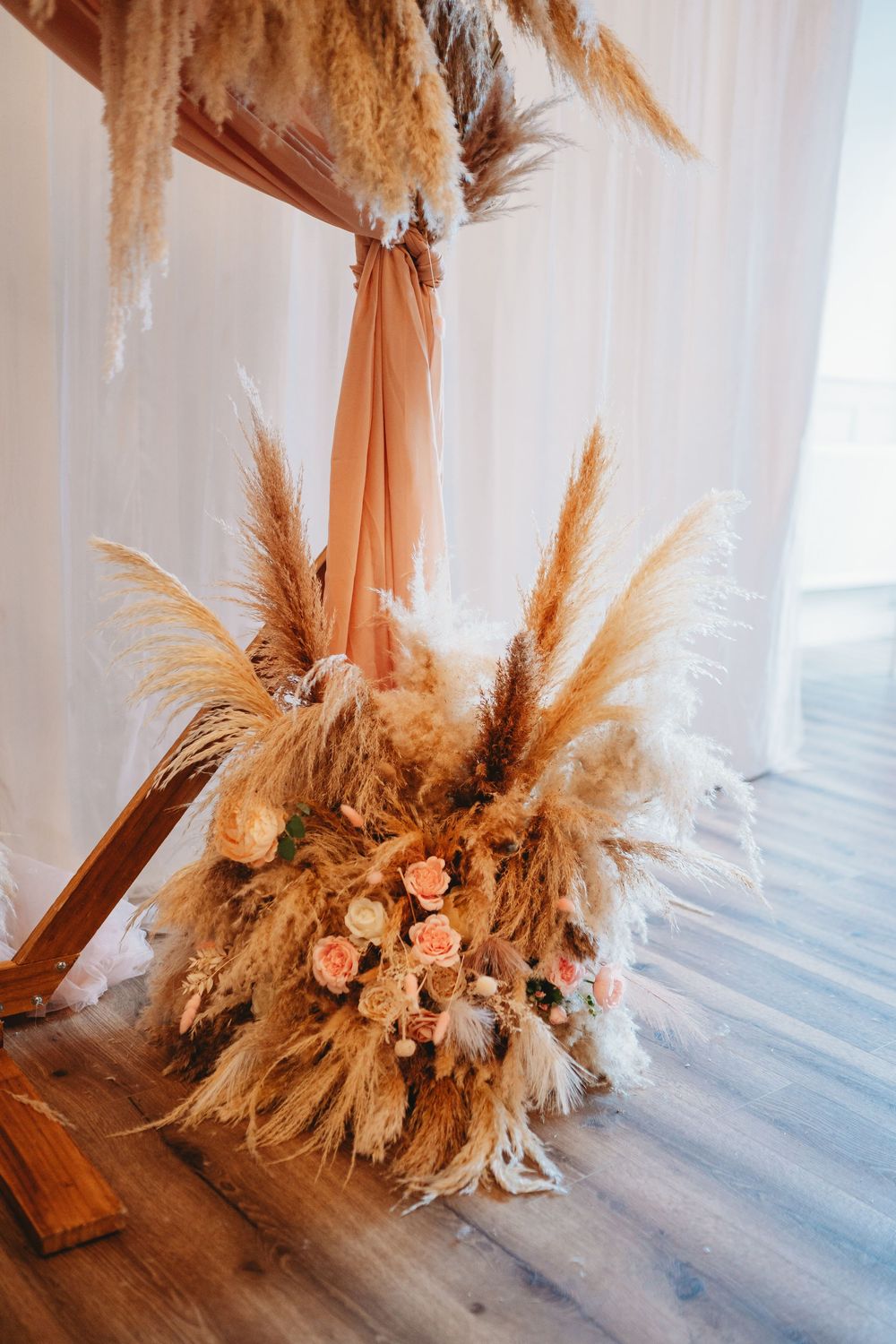 Boho wedding arch decorated with pampas grass and peach roses against white drape backdrop.