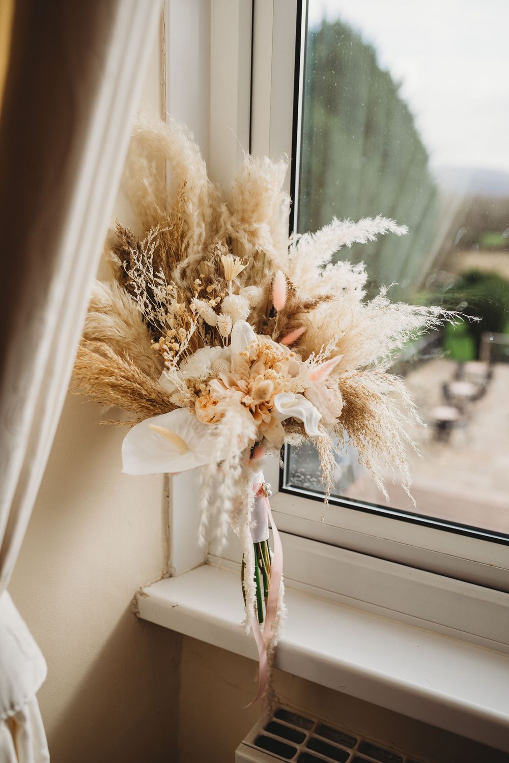 Boho dried flower wedding bouquet with pampas grass and neutral tones resting on a windowsill.