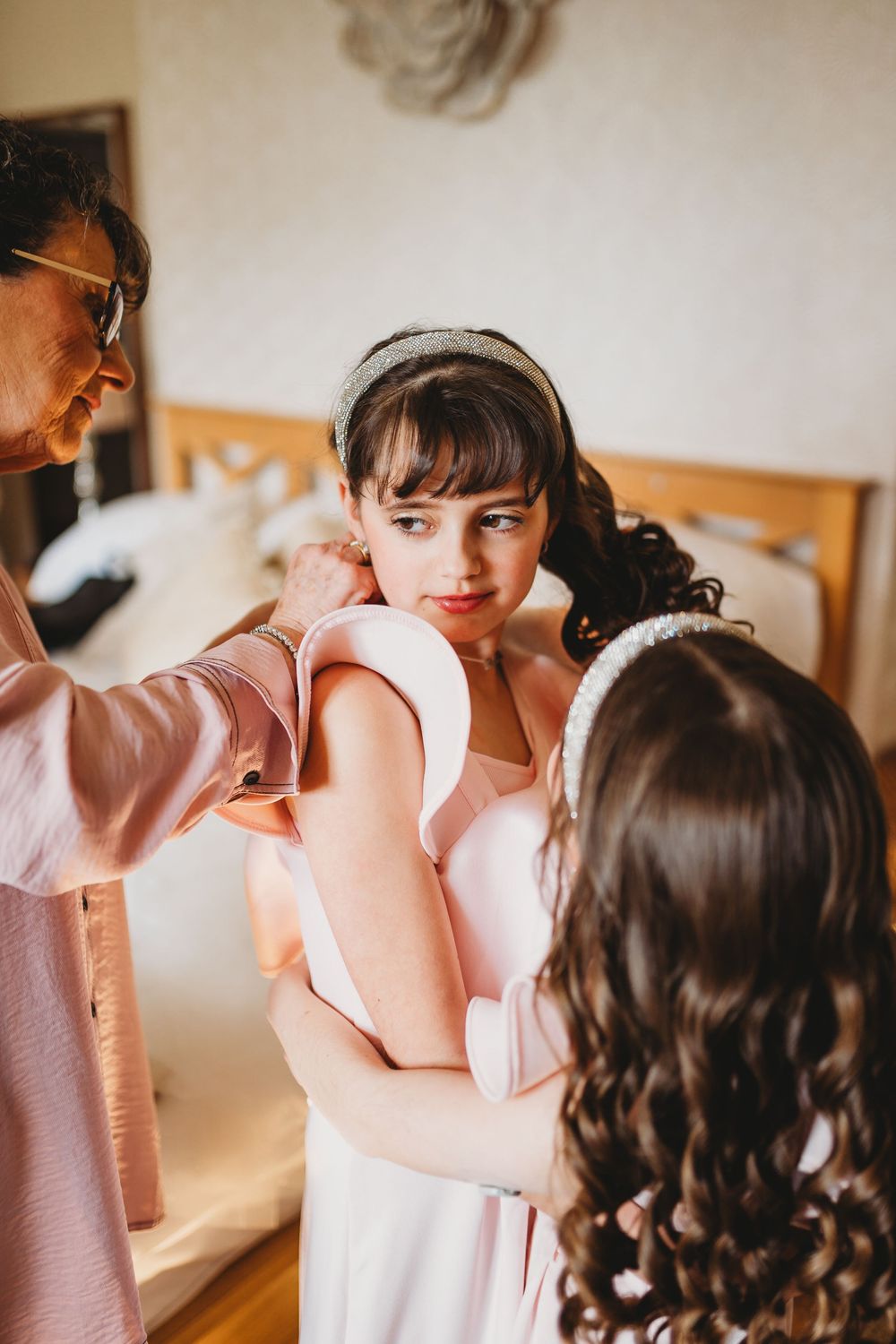 Young flower girl being helped into her pink dress by two women during wedding preparations.