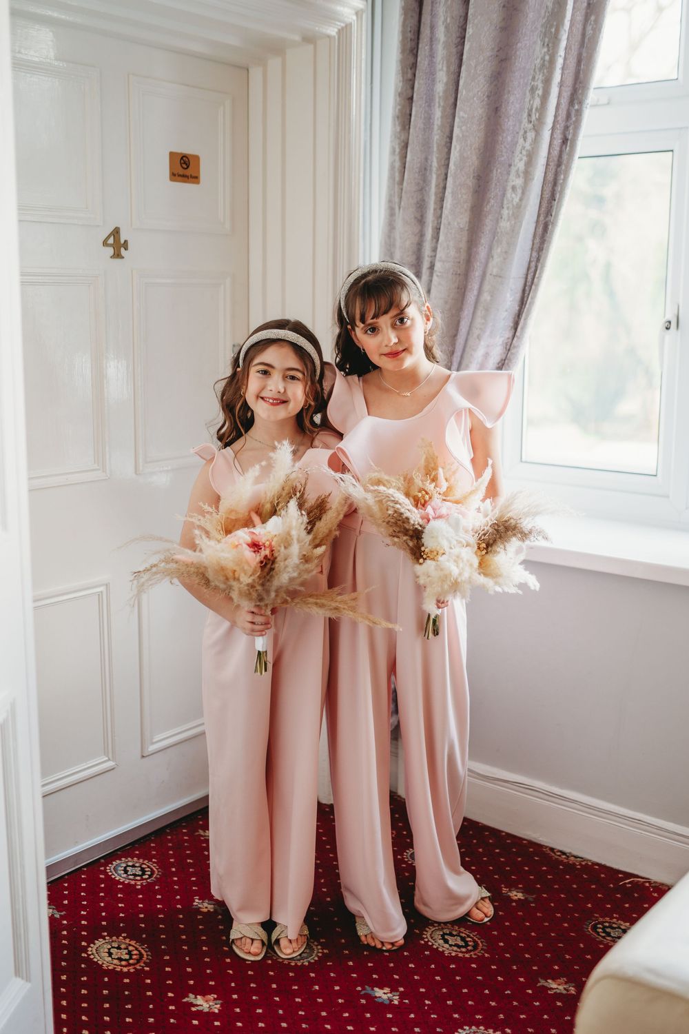 Two young bridesmaids in pink jumpsuits holding boho pampas grass bouquets by a window.