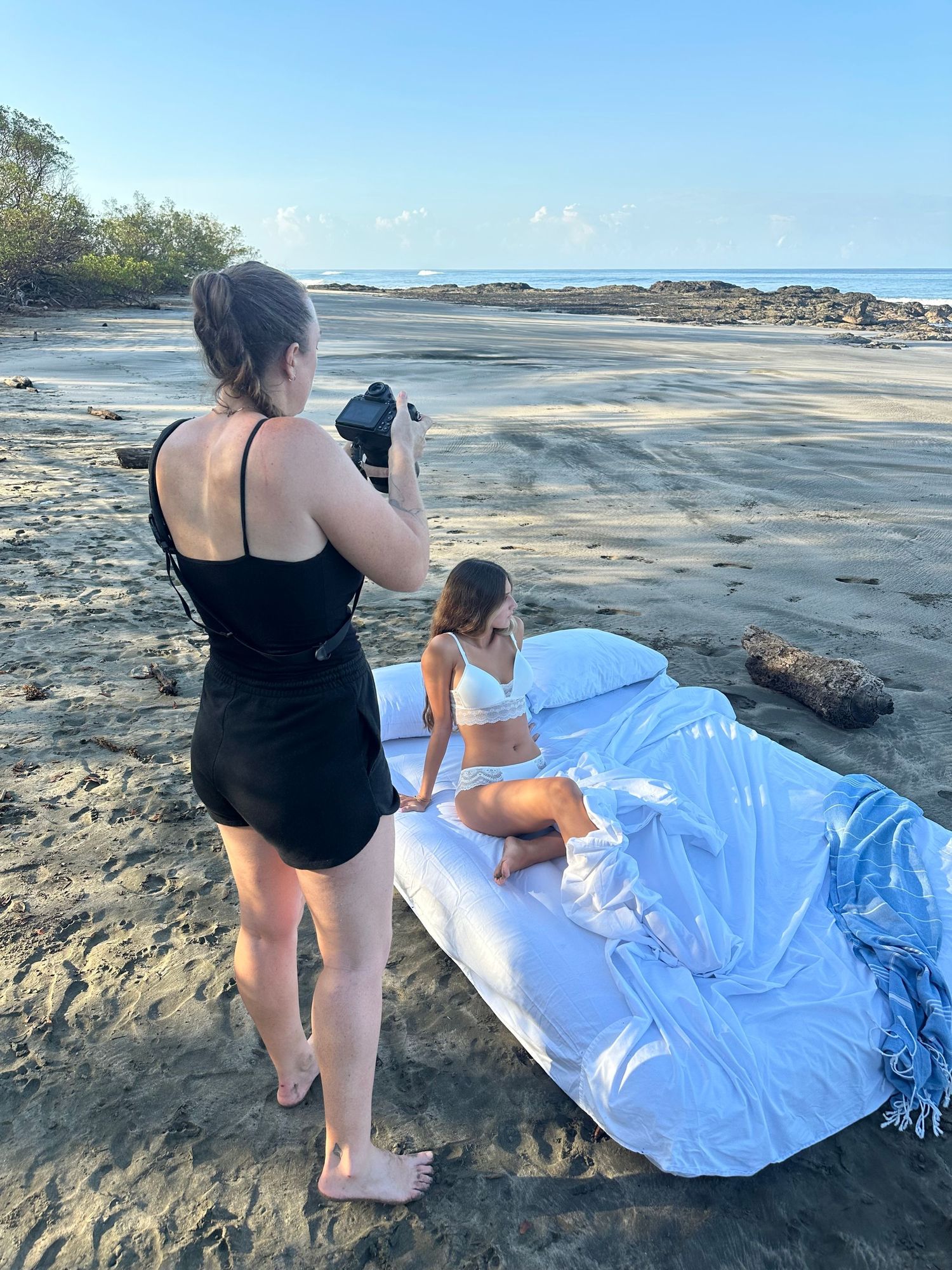 Photographer in black swimsuit taking photos of a model in white bikini on white sheets at a tropical beach.