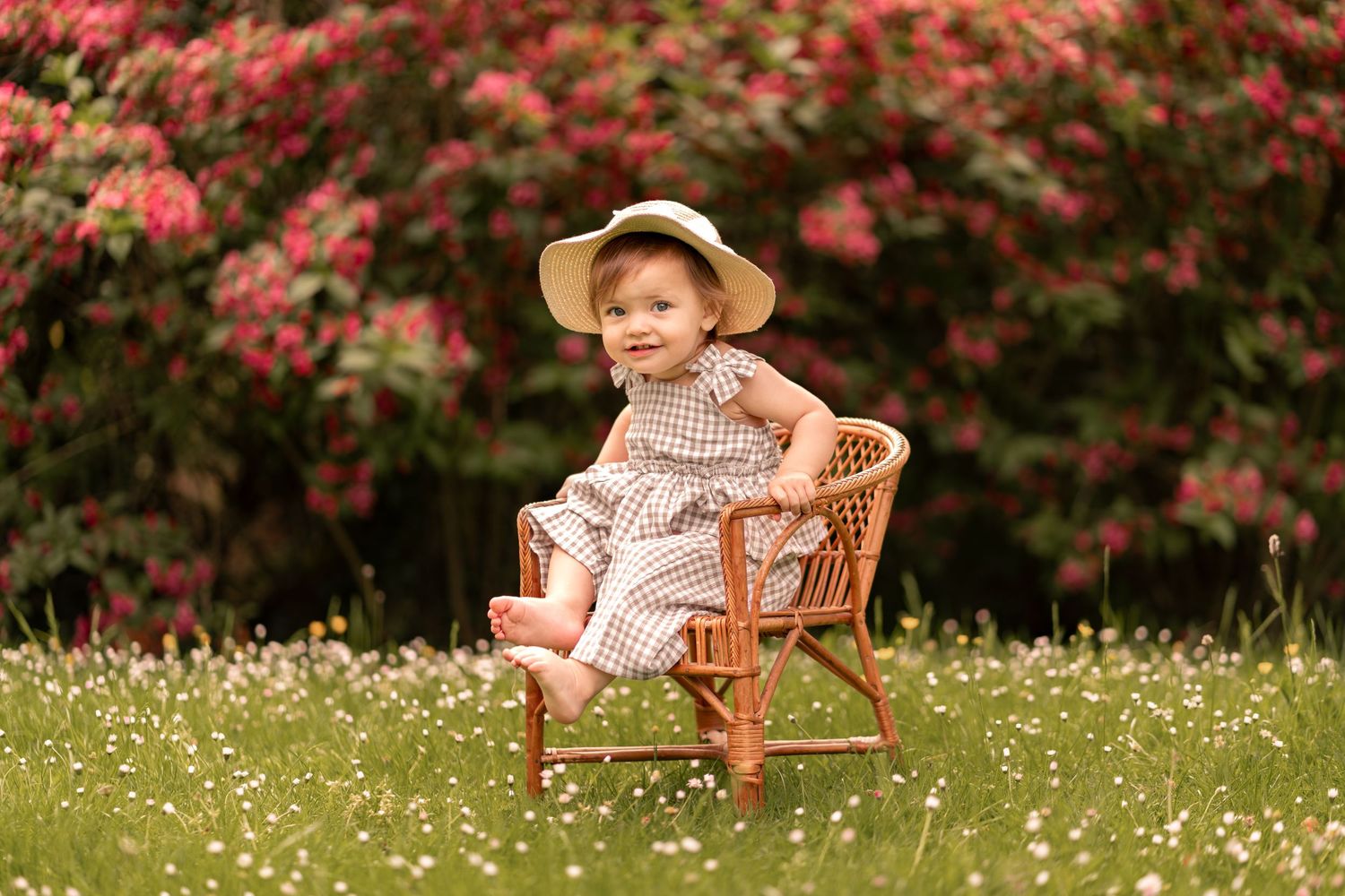 A cute toddler in a plaid dress and straw hat sits on a wicker chair in a blooming garden.