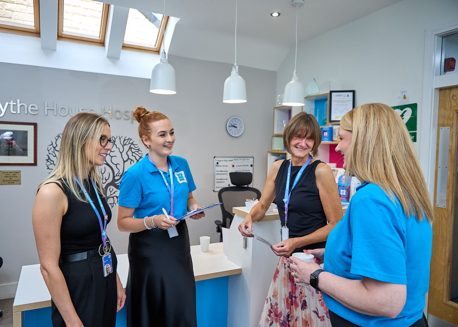 Four women in blue uniforms chat in a bright modern healthcare reception area with pendant lights.