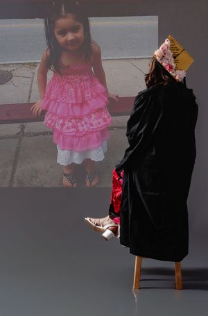 Graduate in black gown and decorated cap sits before projected childhood photo of young girl in pink dress.