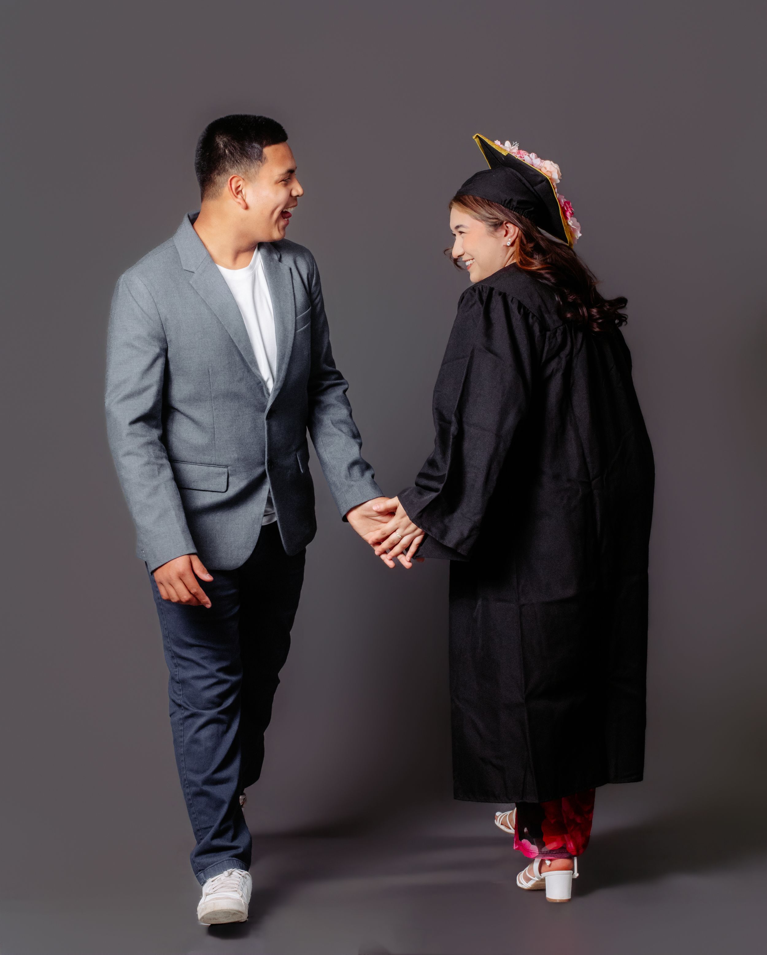 A young man in a gray blazer holds hands with a smiling graduate in cap and gown.