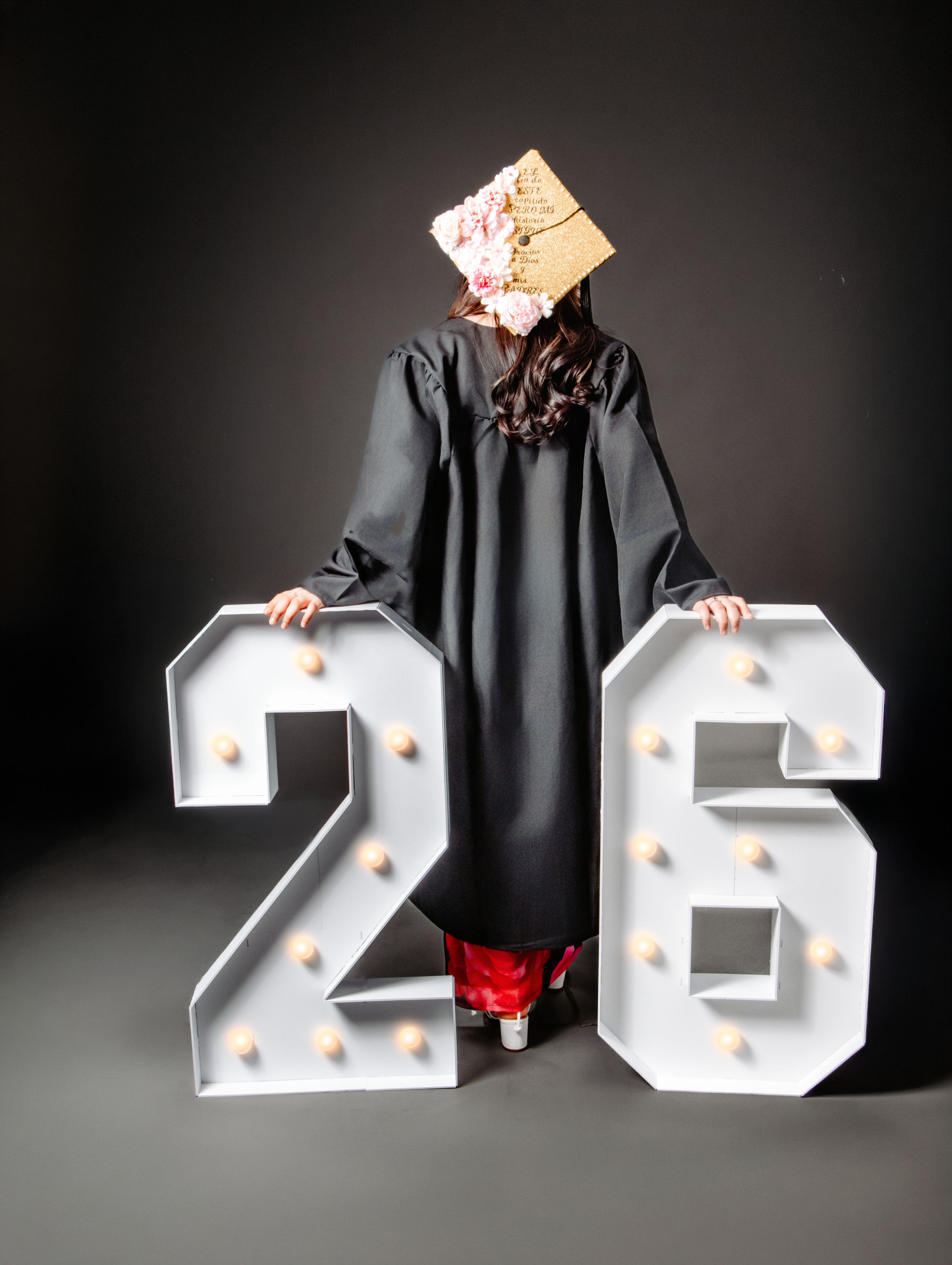 Graduate in black gown holds illuminated marquee numbers 26, wearing decorated cap, celebrating class of 2026.