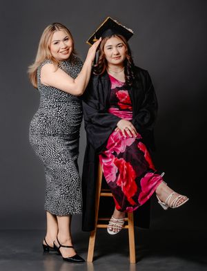 Two women pose for a graduation photo, one in a floral dress wearing a cap and gown, the other in a black dress.