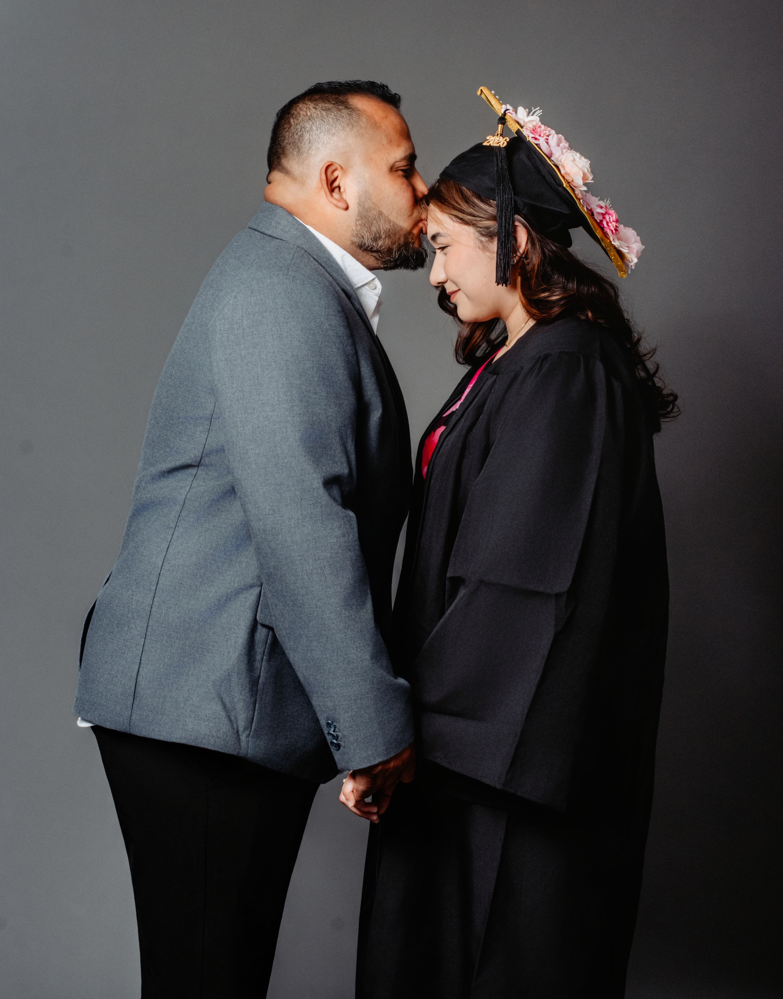 A man in a gray suit kisses a woman wearing a graduation cap and gown while holding hands against a gray background.