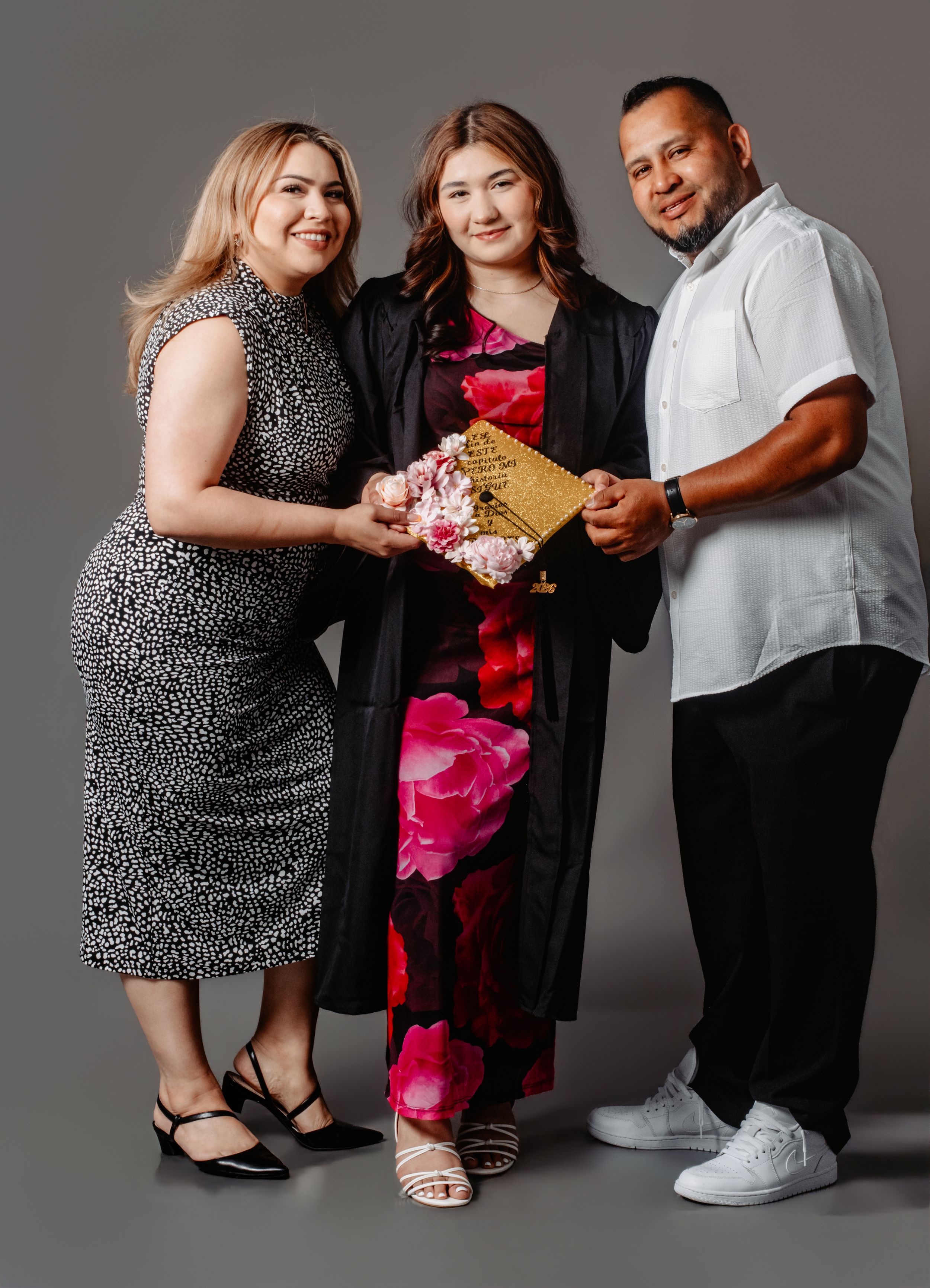 A graduate in cap and gown poses with two proud family members holding flowers and a gold clutch.