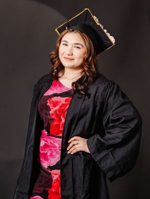 Young woman in black graduation cap and gown over floral dress poses confidently for graduation portrait.