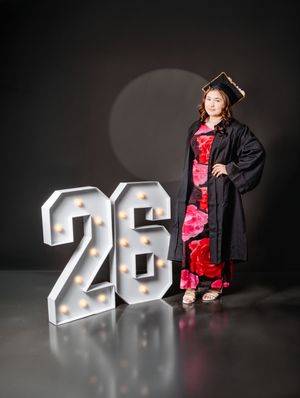 Graduate in black cap and gown with floral dress poses beside illuminated marquee number 26 on dark studio background.