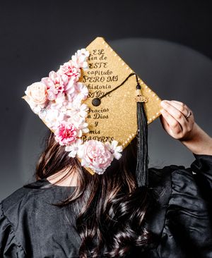 Gold glitter graduation cap decorated with pink flowers and Spanish text thanking God and parents, worn by graduate in black gown.