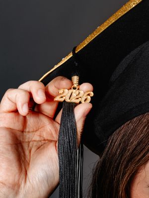 A close-up of a 2026 gold tassel charm on a black and gold graduation cap held by a graduate.
