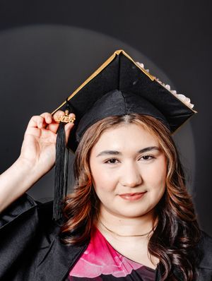A young woman in a black graduation cap and gown holds her tassel while smiling at the camera.