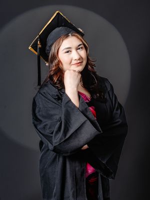 A young woman in black graduation cap and gown poses confidently against a dark studio background.
