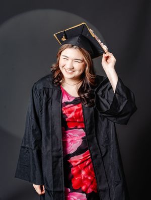 Smiling graduate in black cap and gown over floral dress, posing for graduation portrait photo.