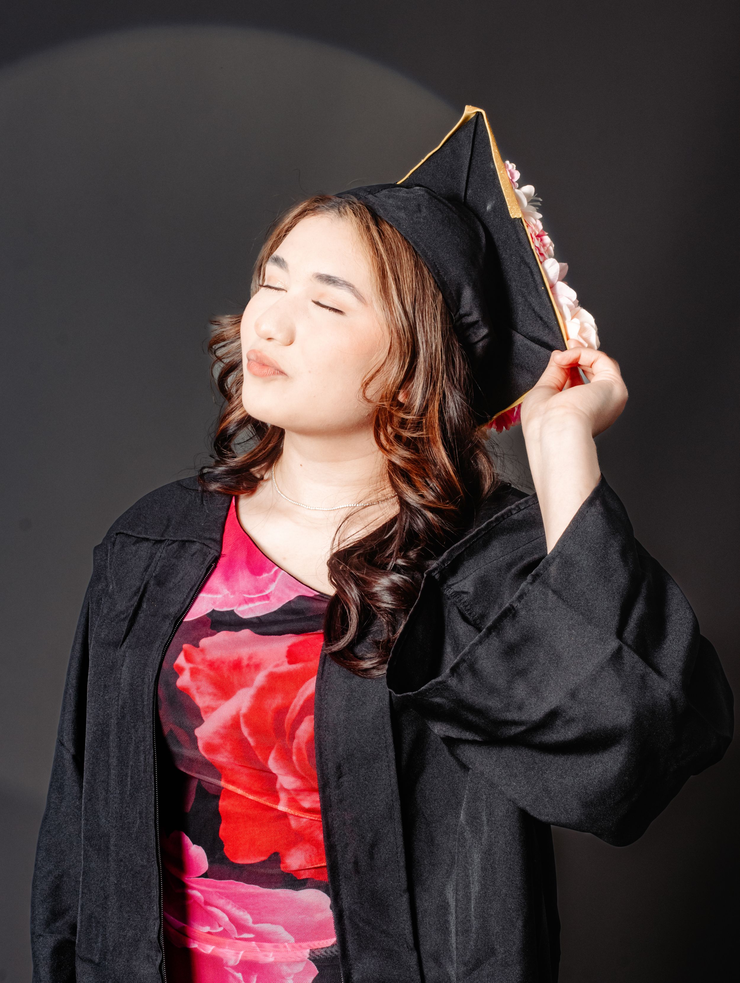 Young woman in black graduation cap and gown posing confidently with eyes closed against dark background.