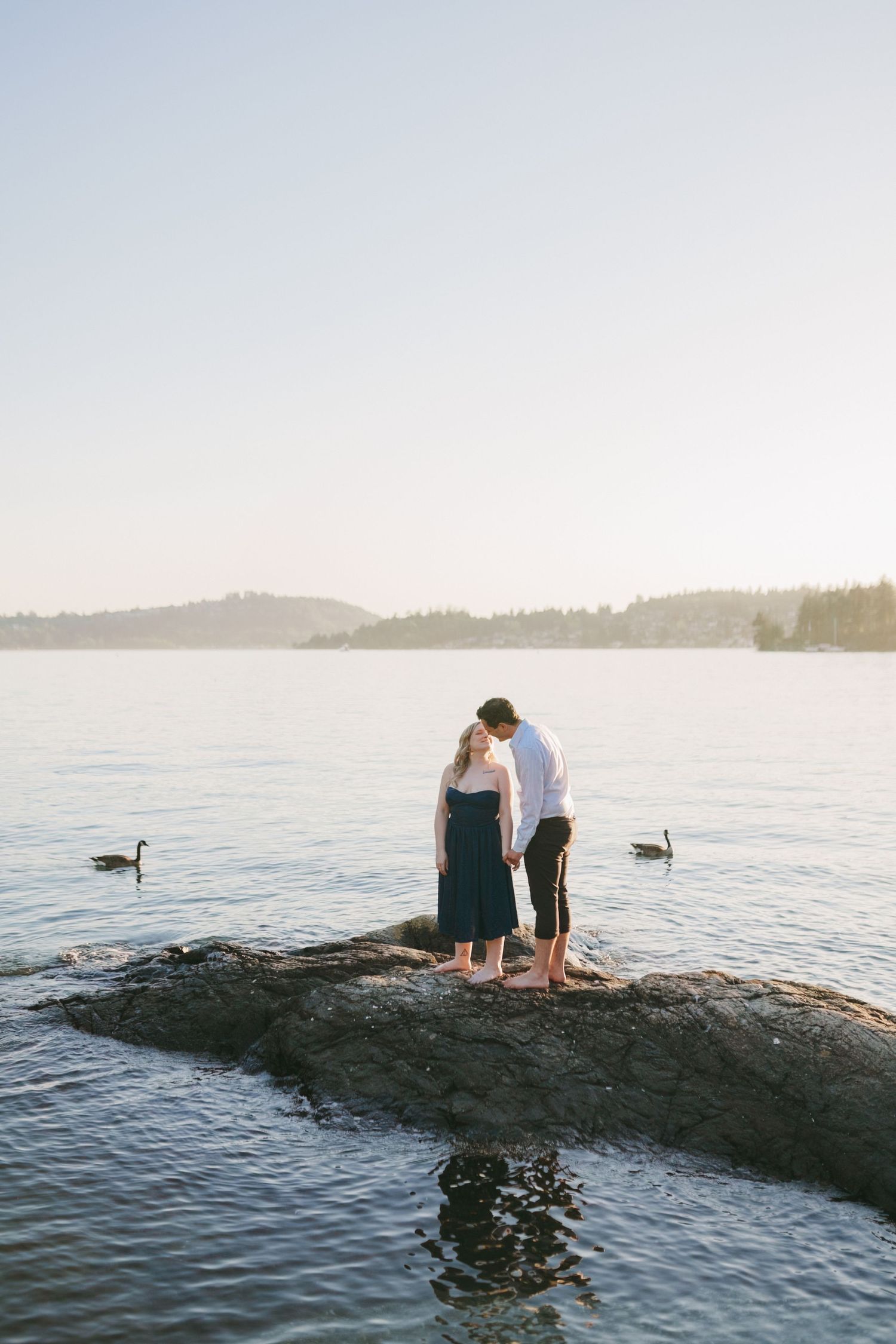 Couple standing barefoot on rocky shoreline at sunset, holding hands with geese swimming nearby and misty trees in background.