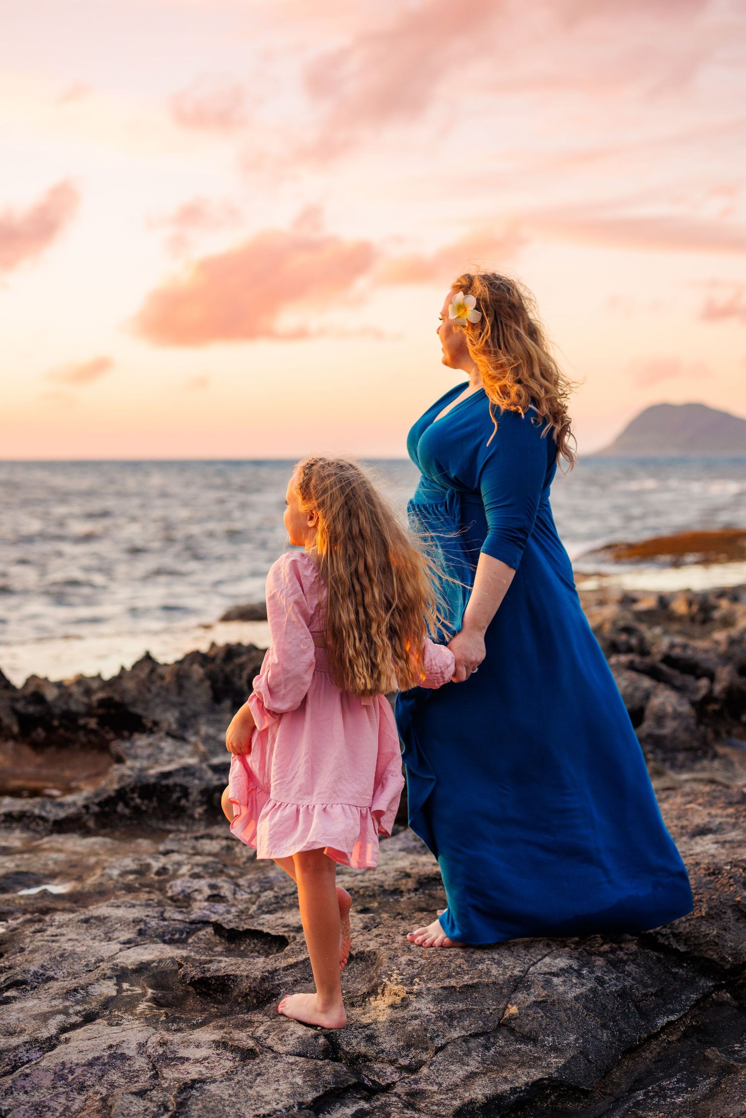 A mother and daughter in flowing dresses hold hands on rocky shore watching a vibrant pink sunset over the ocean.