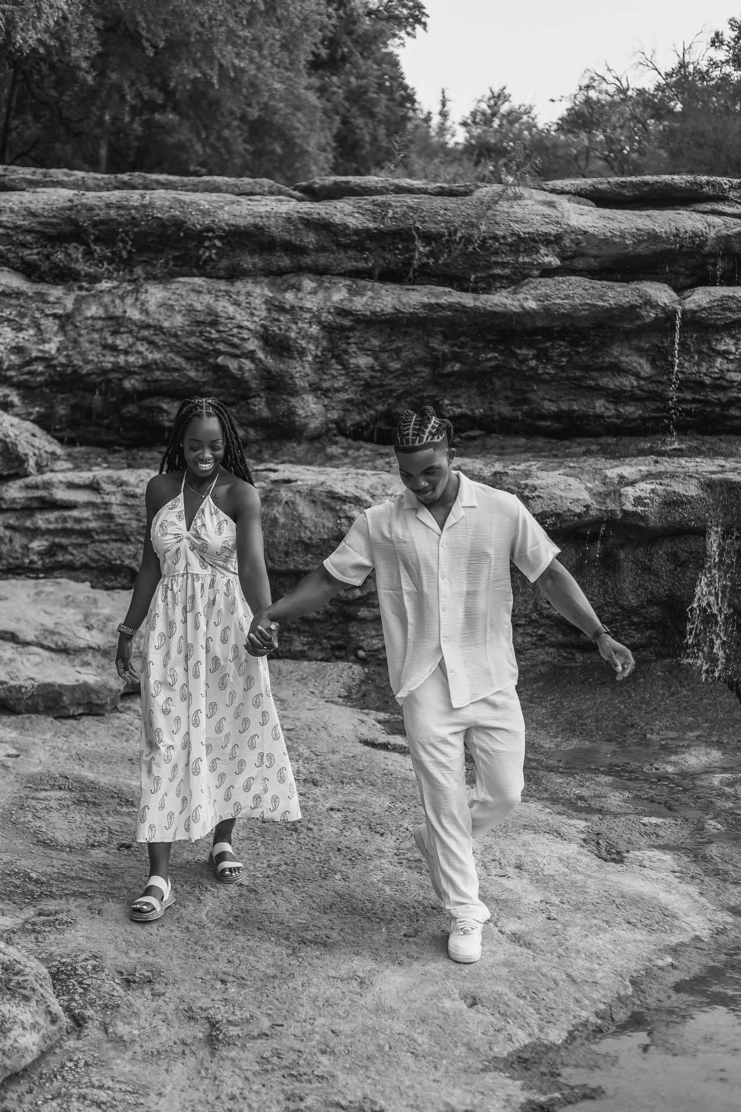 Black and white photo of a couple holding hands walking on rocky terrain with trees and stone cliffs in the background.