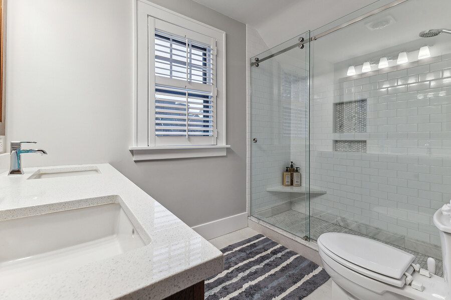 Modern white bathroom with glass walk-in shower, marble countertop vanity, and natural light from window.
