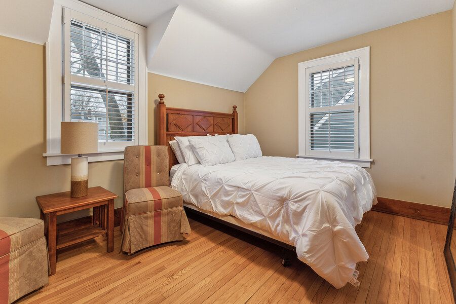 Cozy bedroom with vaulted ceiling, hardwood floors, queen bed with white bedding, striped armchair, and natural light from multiple windows.