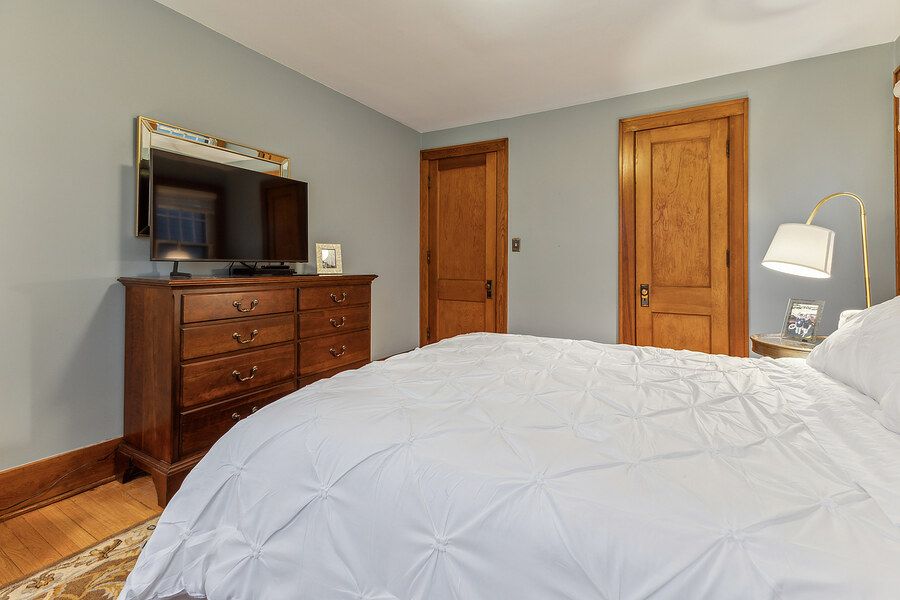 Cozy bedroom with white bedding, wooden dresser with wall-mounted TV, and natural wood doors in a softly painted room.