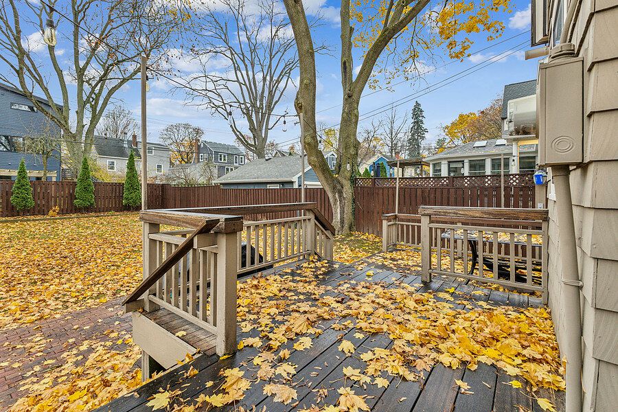 Wooden deck covered in yellow autumn leaves surrounded by tall trees and a privacy fence in a residential backyard.
