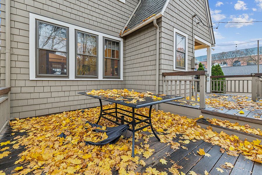 Backyard deck covered in autumn leaves with patio table set against gray shingle siding home.