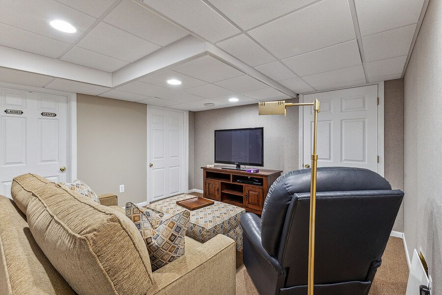 Finished basement rec room with drop ceiling, sectional sofa, leather recliner, flat-screen TV, and neutral carpet flooring.