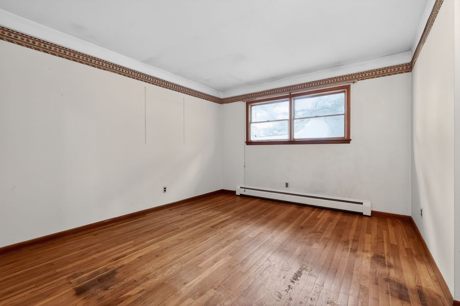 Empty room with hardwood floors, white walls, decorative border trim, and a single window with baseboard heater.