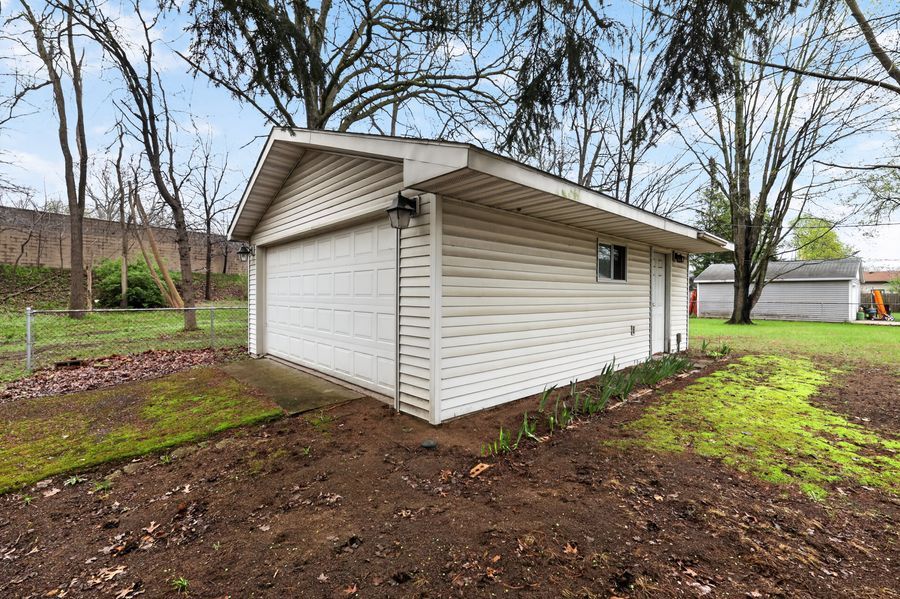 White vinyl detached garage with overhead door sits on a dirt lot surrounded by green grass and bare trees.