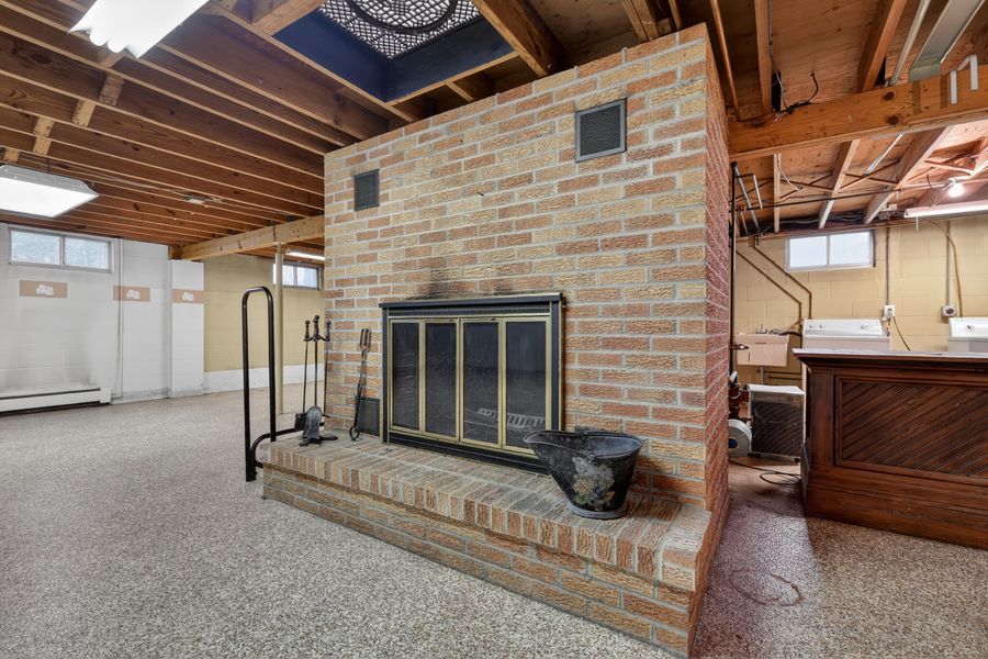 Unfinished basement with brick fireplace, glass doors, fireplace tools, exposed wood beam ceiling, and carpeted floors.