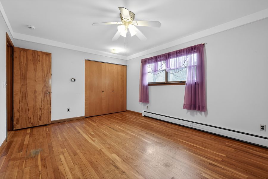 Bright bedroom with hardwood floors, ceiling fan, crown molding, baseboard heater, and double closet with purple curtains.
