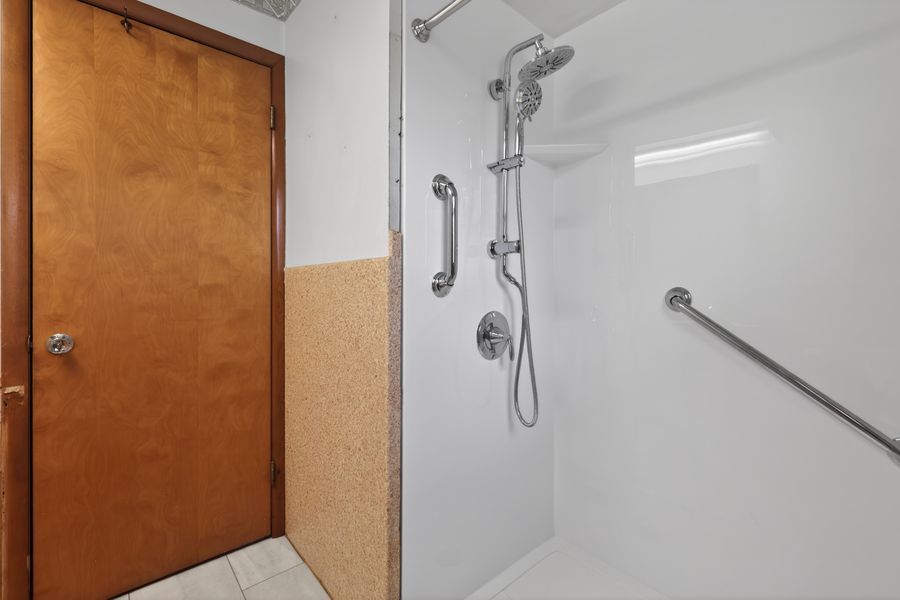 Accessible walk-in shower with grab bars, handheld showerhead, and wooden door in a clean white bathroom.