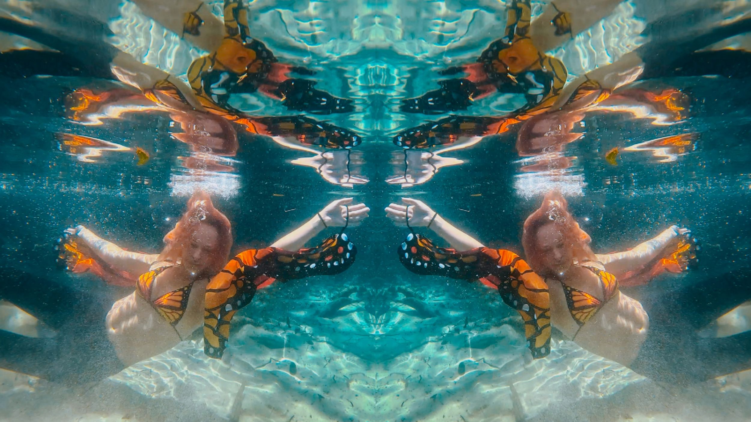 Mirrored underwater photo of swimmers with colorful orange floral accessories in crystal clear blue water.