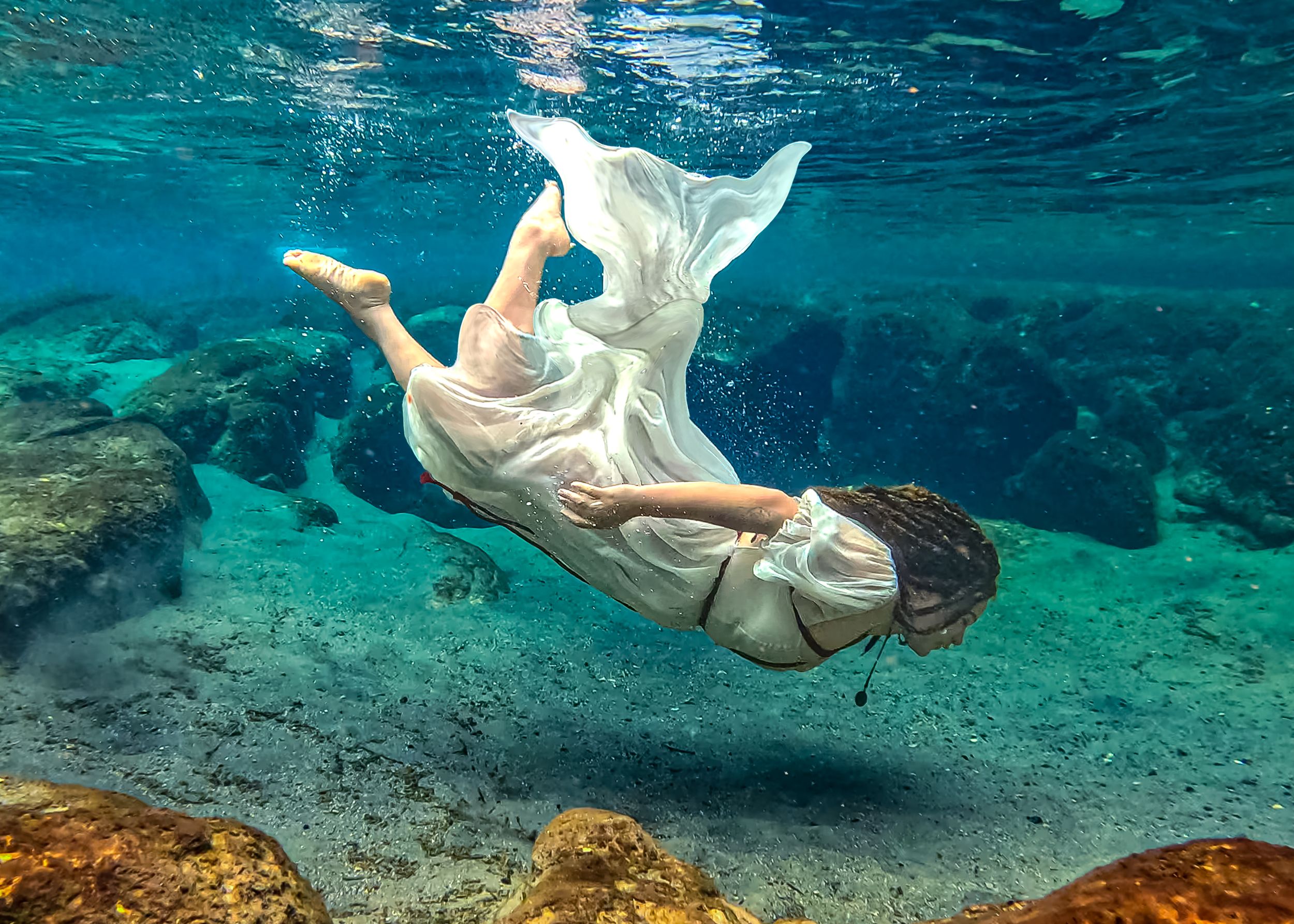 Woman in white dress gracefully swimming underwater among rocky ocean floor in clear turquoise water.