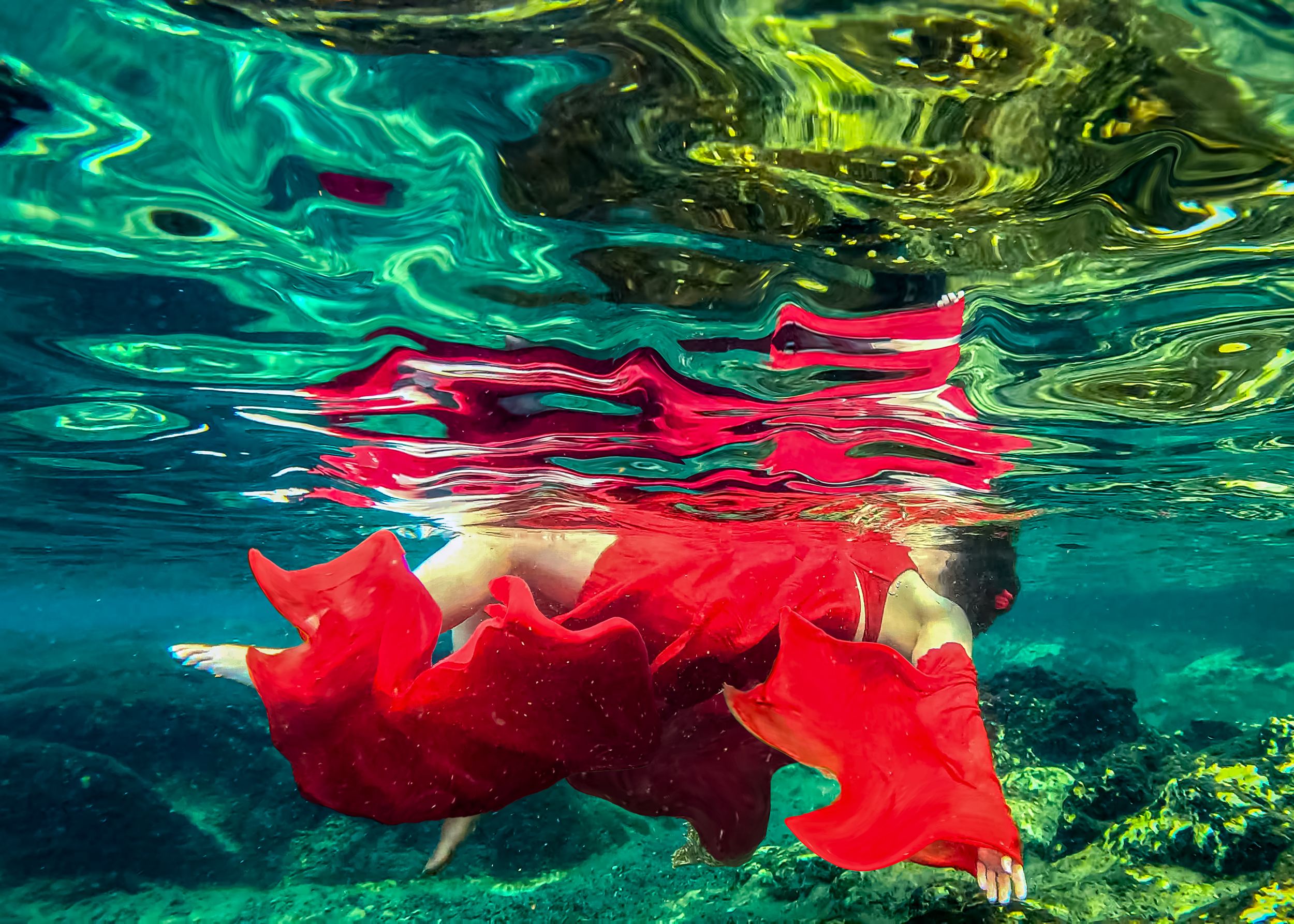 Woman in flowing red dress swimming underwater in crystal clear turquoise water with colorful reflections on surface.