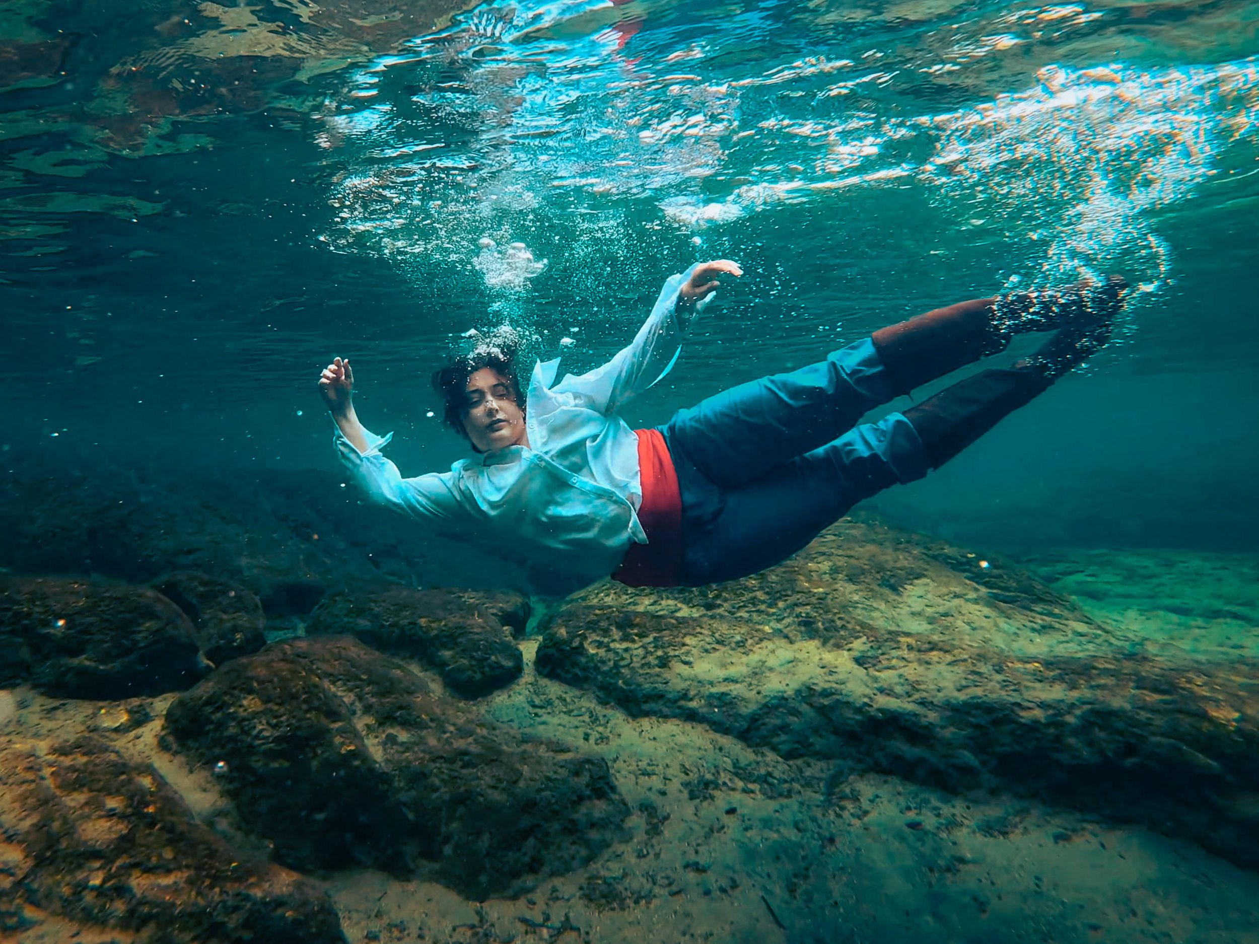 Person swimming underwater near rocks in clear turquoise ocean water, wearing casual clothes and snorkeling gear.