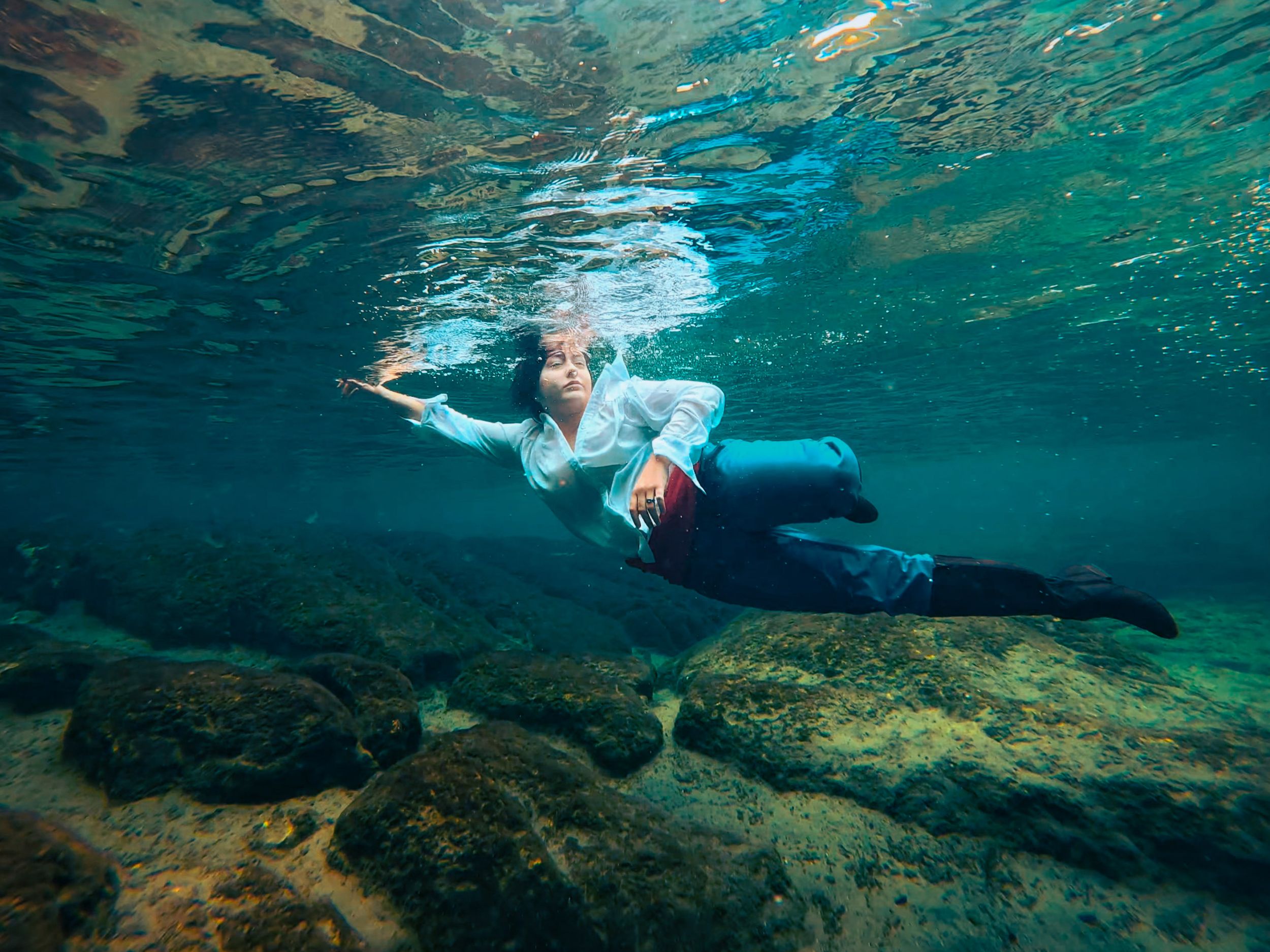 Person swimming underwater in clear turquoise water above rocky bottom, wearing white shirt and dark pants.