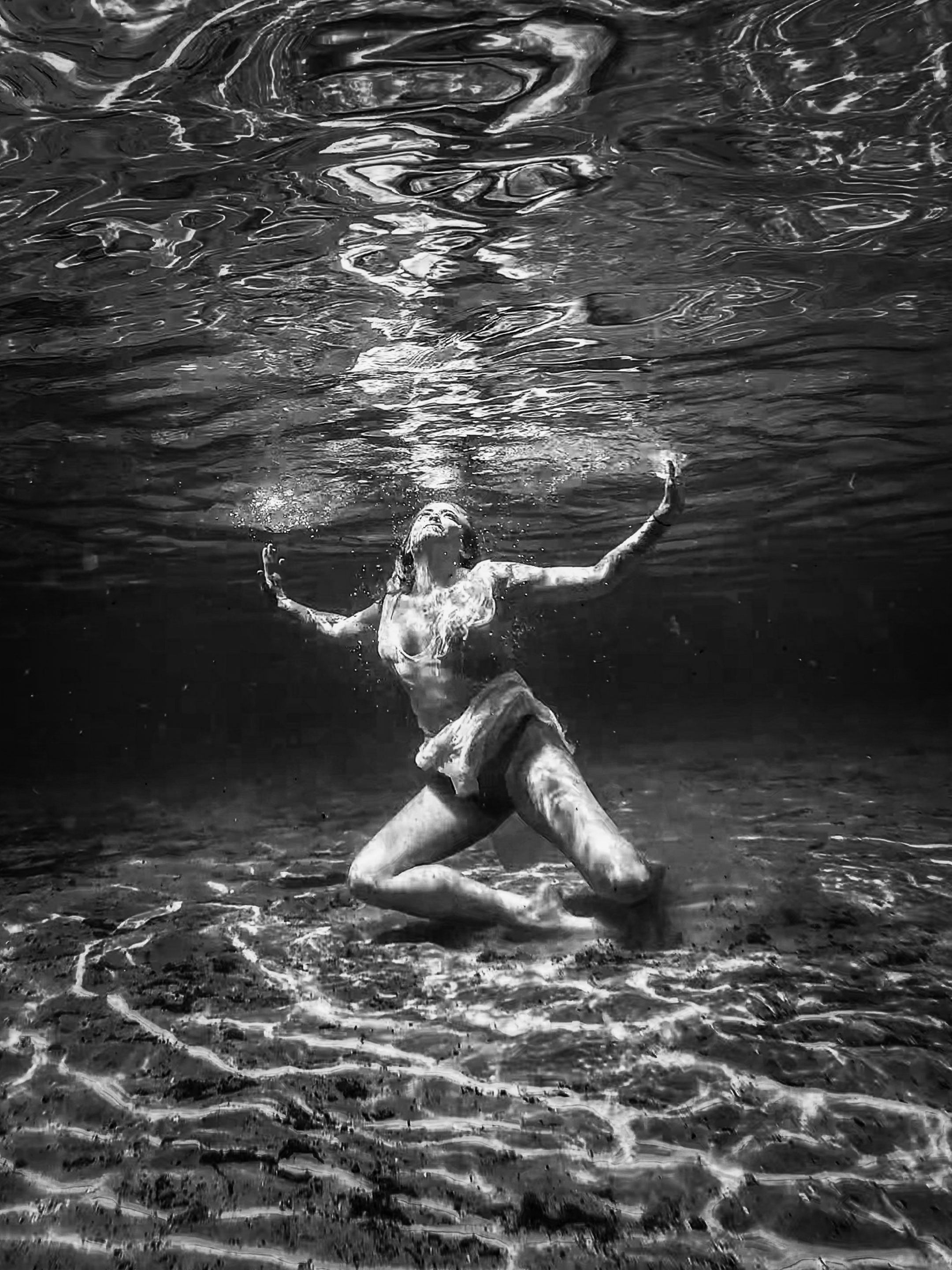 Black and white underwater photo of a woman in a bikini gracefully posing on the sandy bottom.