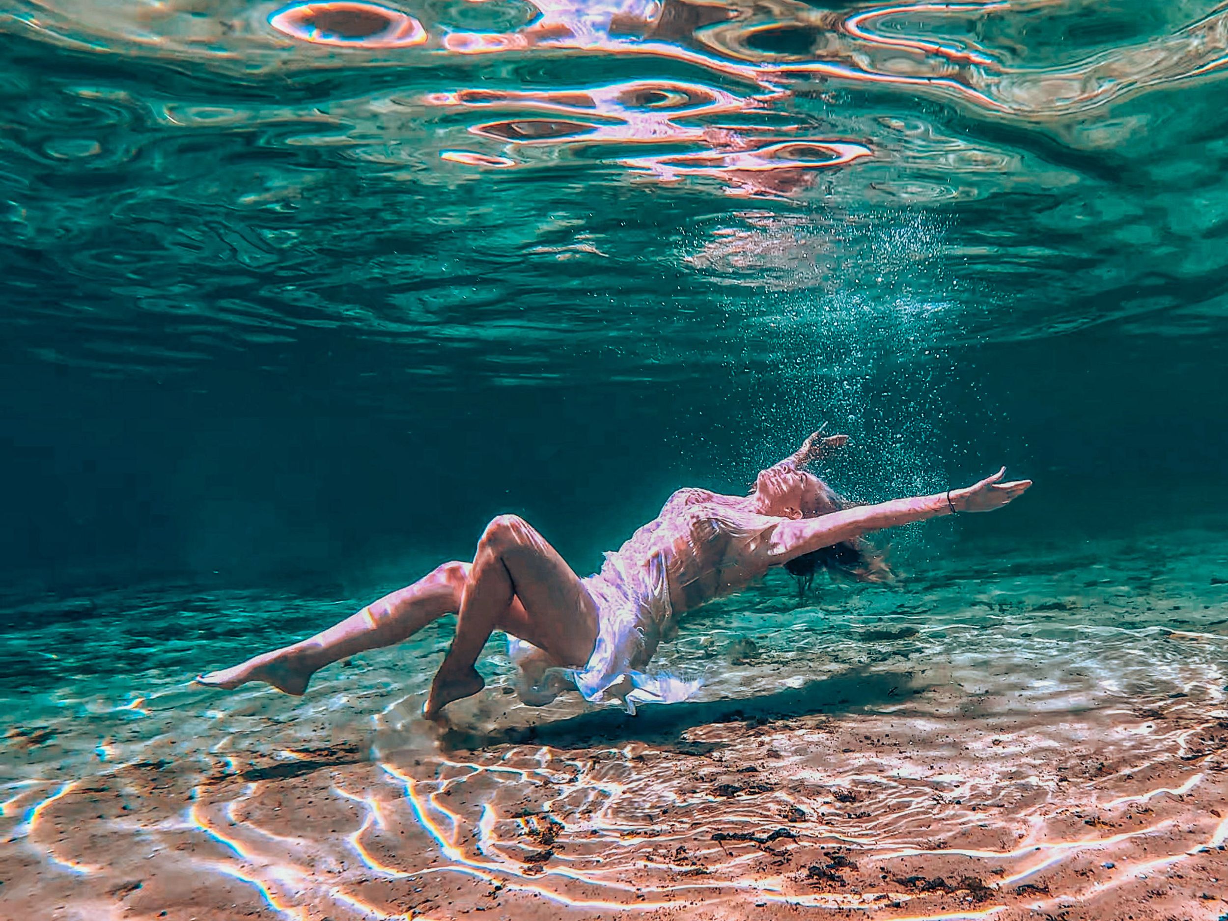 Woman in white dress floating gracefully underwater in clear turquoise water with sandy bottom and sunlight filtering through.