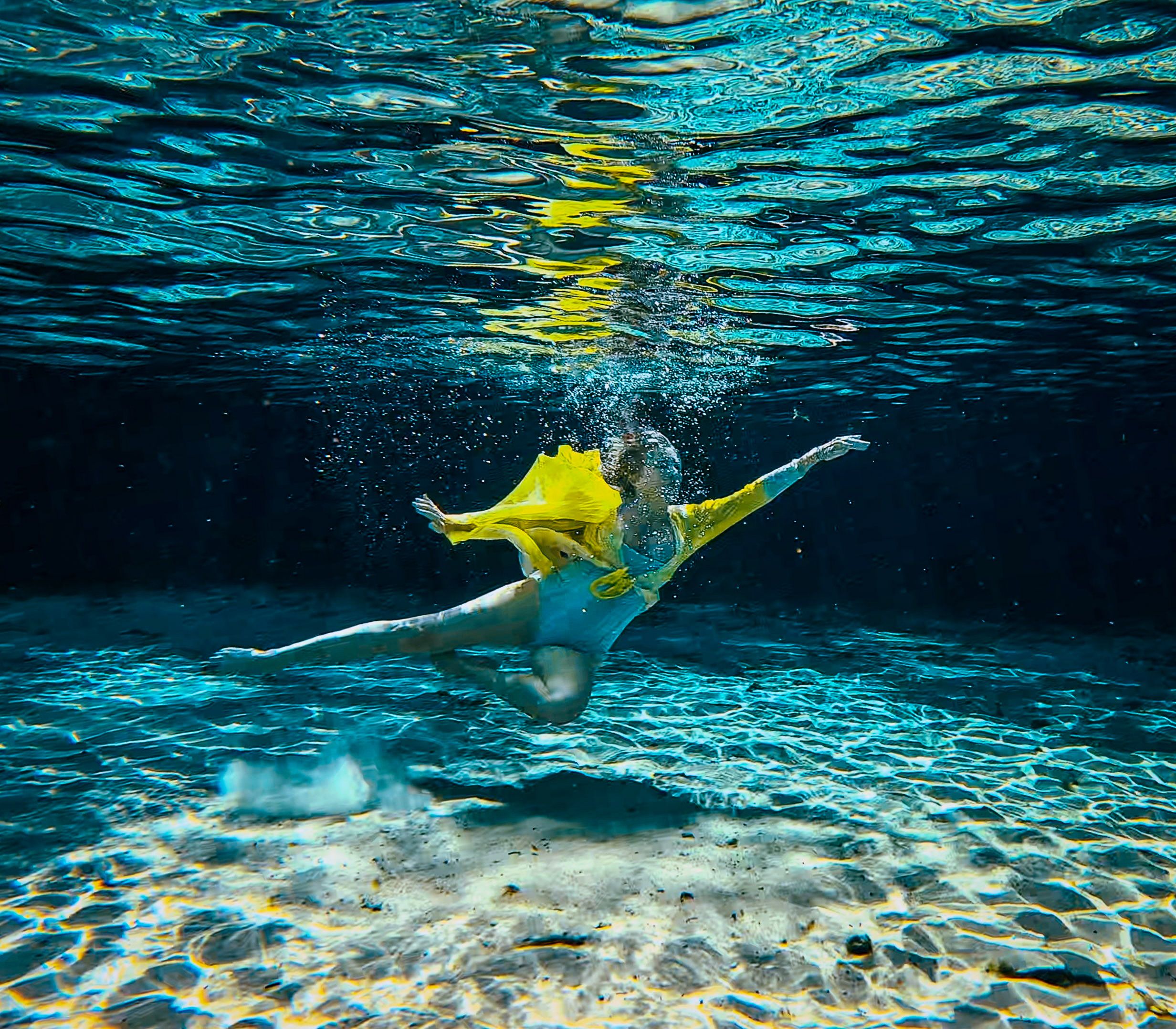 Graceful dancer in yellow and blue dress performing underwater ballet in a clear blue pool with sunlight filtering through.