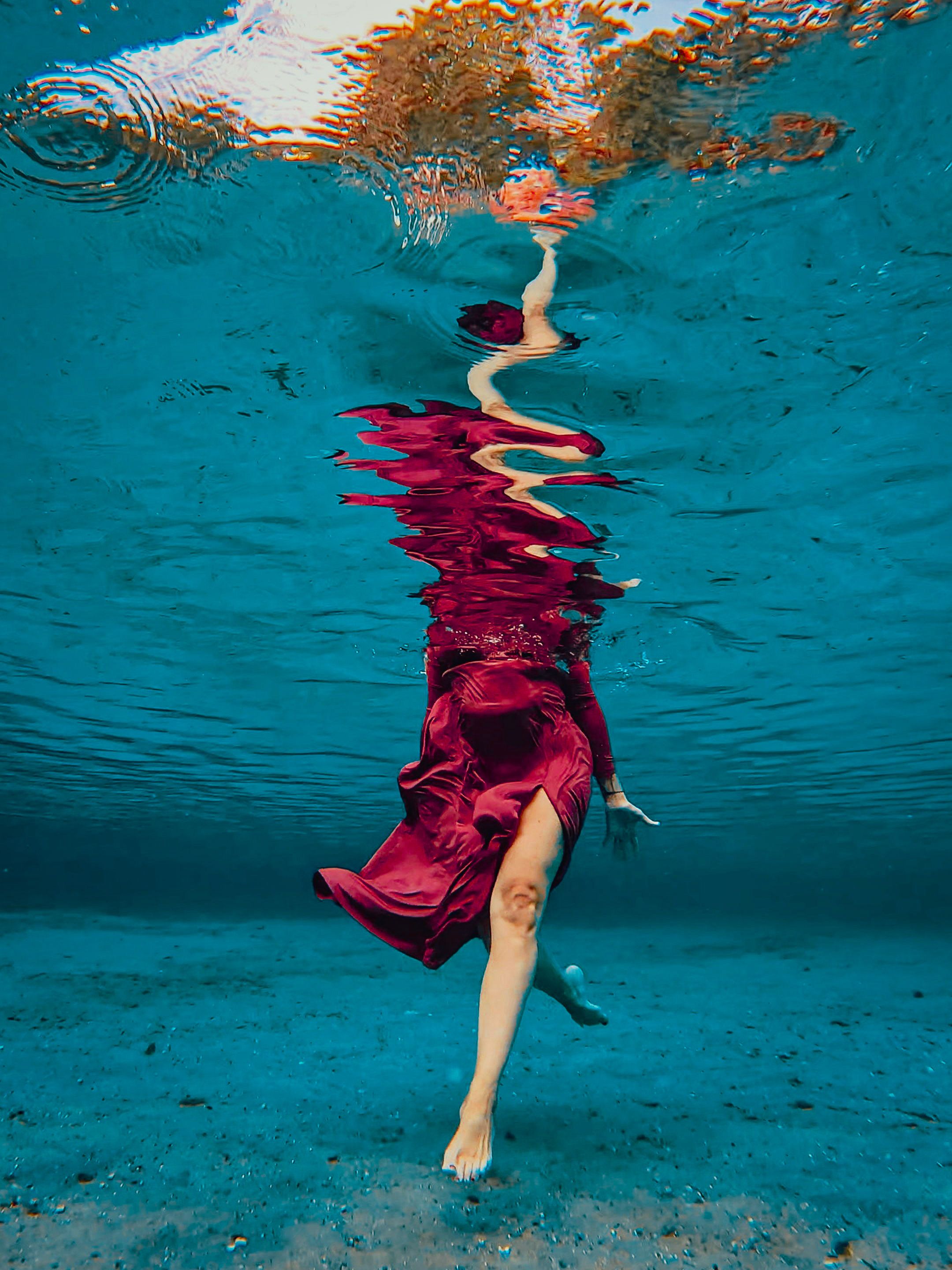 Woman in flowing red dress captured underwater in a turquoise pool, her silhouette elegantly distorted by the water surface.
